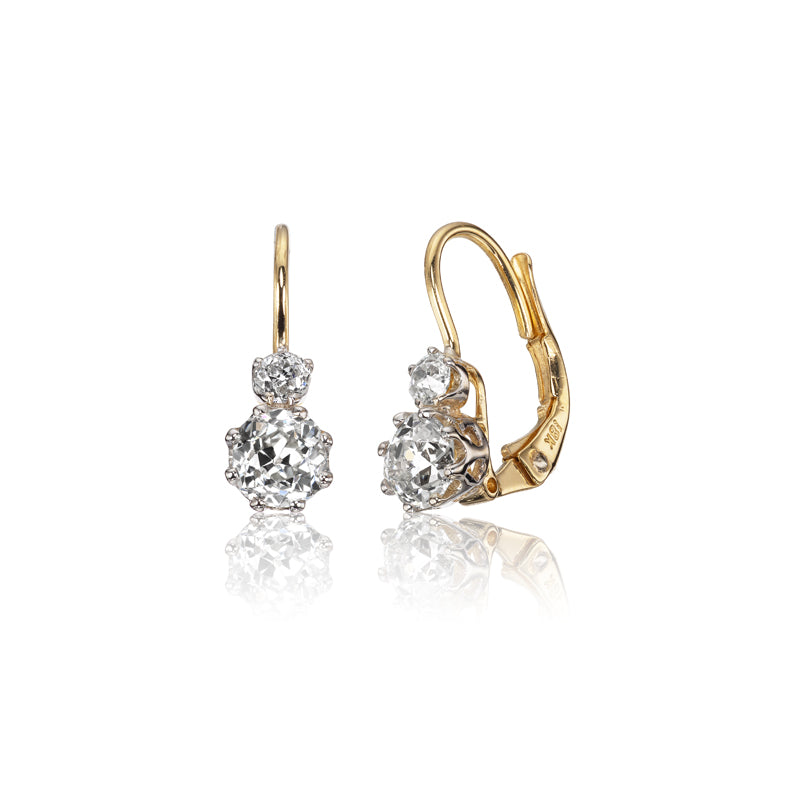 The Estelle 1.29 features gold drop earrings, each with a large round diamond accented by a smaller diamond above, set in prongs with lever-back clasps, displayed on a white background.